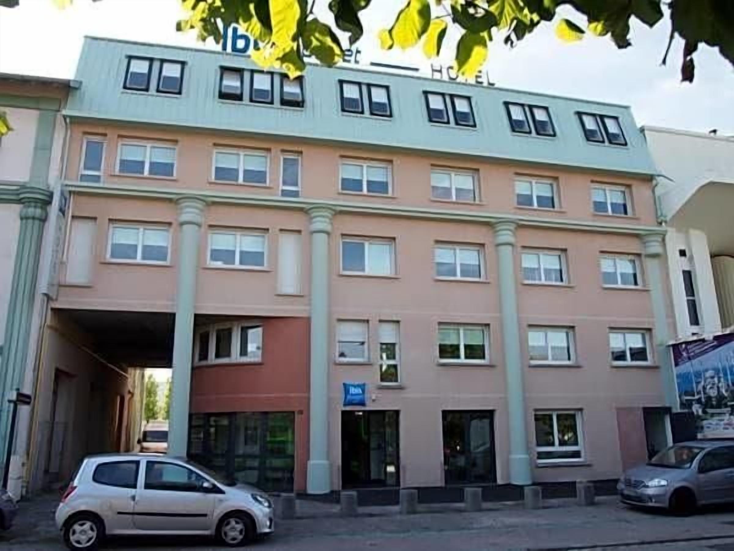 Un hotel de varios pisos con una entrada central, un estacionamiento visible y un letrero que indica una zona de negocios o servicios.