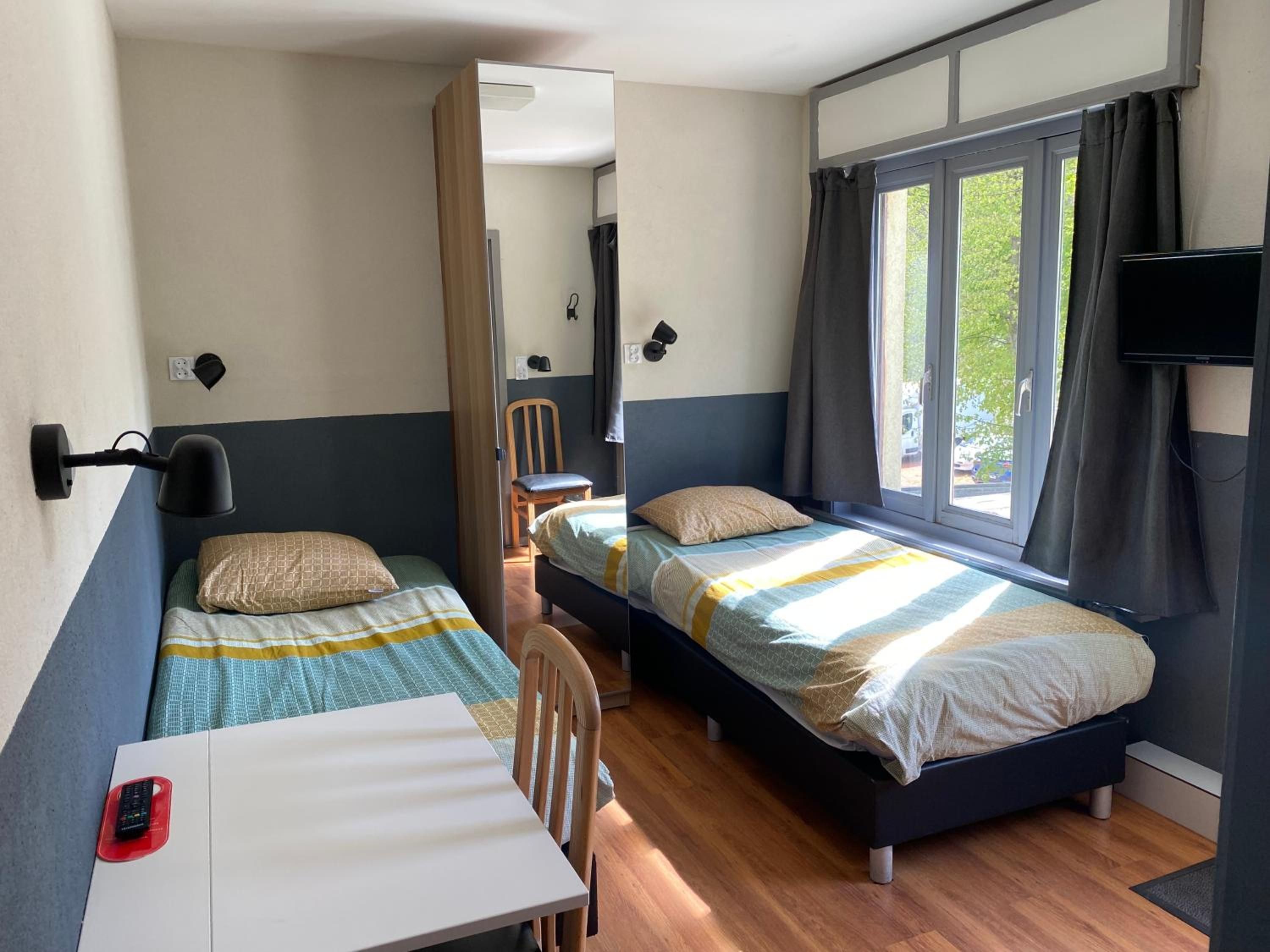 Habitación pequeña con dos camas individuales, una mesa, una silla, televisión y ventana con cortinas.