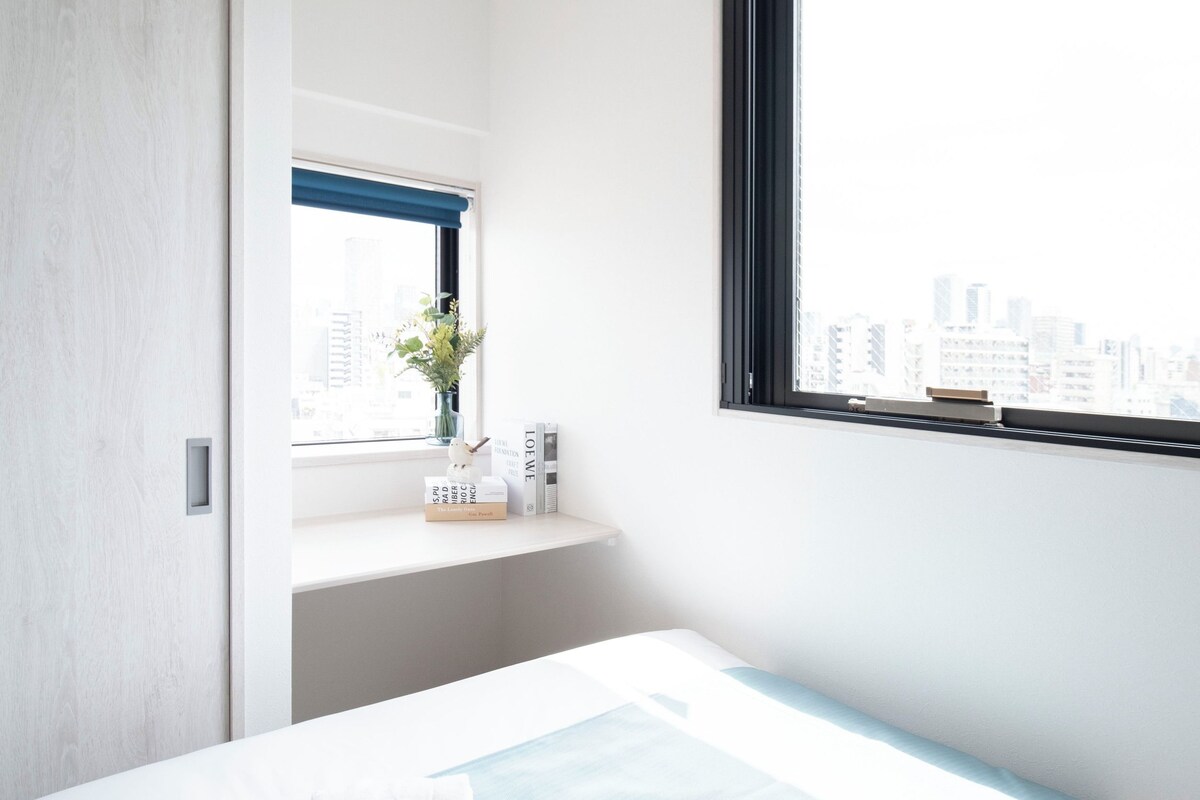 A hotel room with a bed, a window with a view of the city, a bedside table with a vase of flowers, and a door.