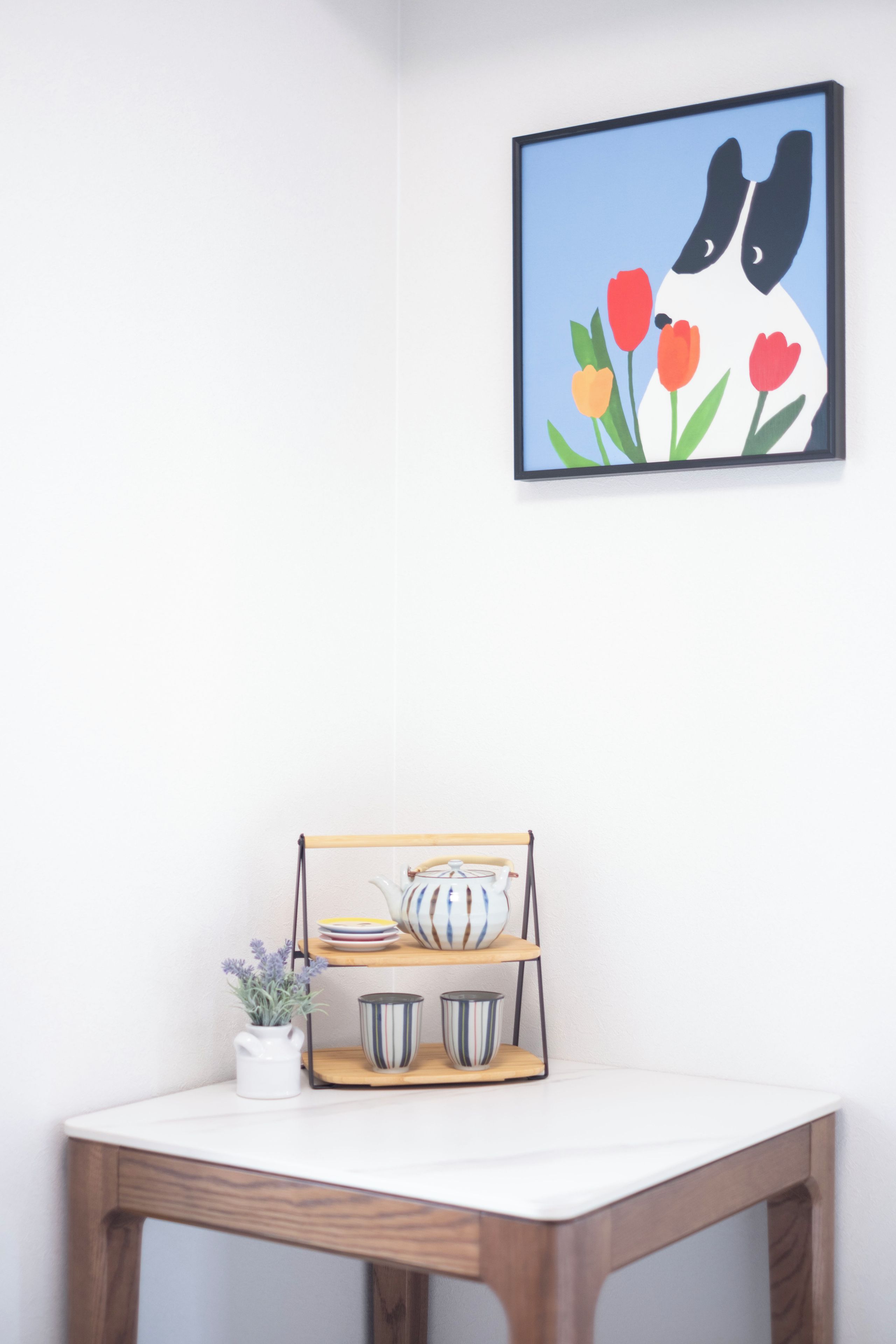 Uma obra de arte emoldurada com pássaros e tulipas numa parede, uma mesa pequena com um tabuleiro contendo chávenas e um bule, e uma planta em vaso.