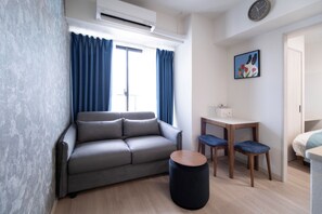 A modern living room with a grey sofa, a round black ottoman, a wooden side table, and a small blue chair. - The Peak Chiyozaki Elite (Chiyozaki, Nishi Ward)