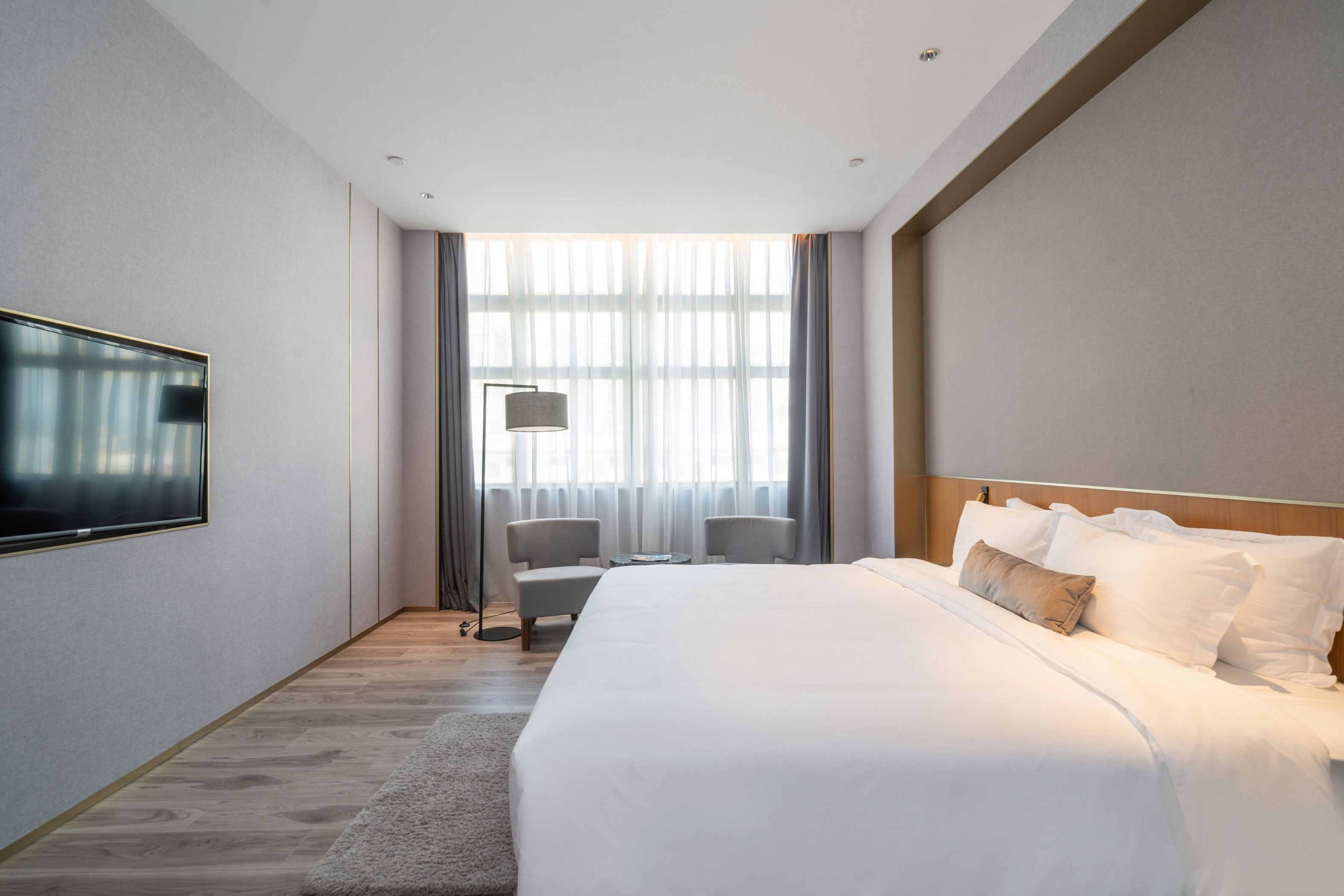 A hotel room with a large bed, a flat-screen TV mounted on the wall, a seating area with a chair, and a large window with curtains.