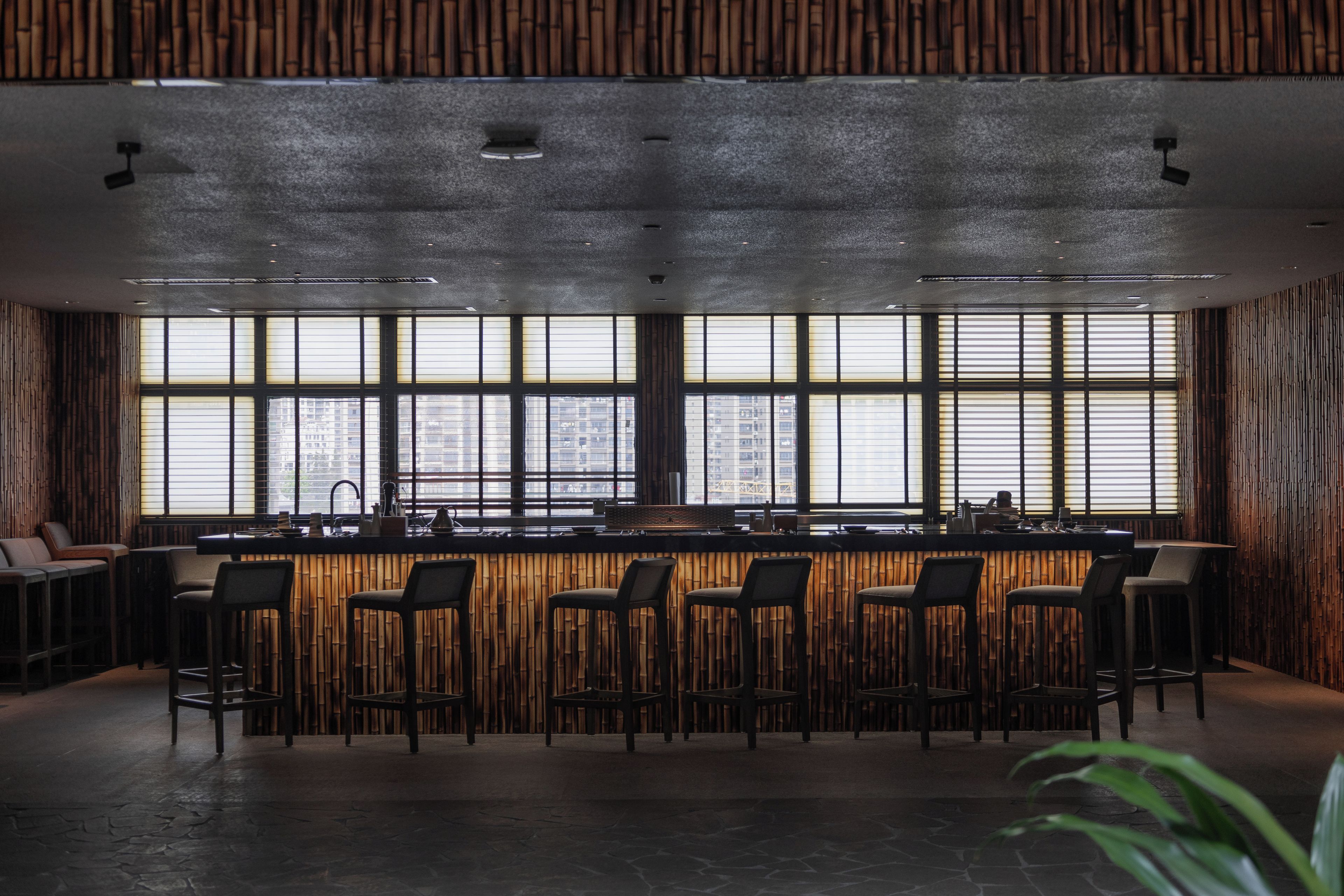 A modern bar area with a long counter, multiple stools, and large windows providing a view of the cityscape.