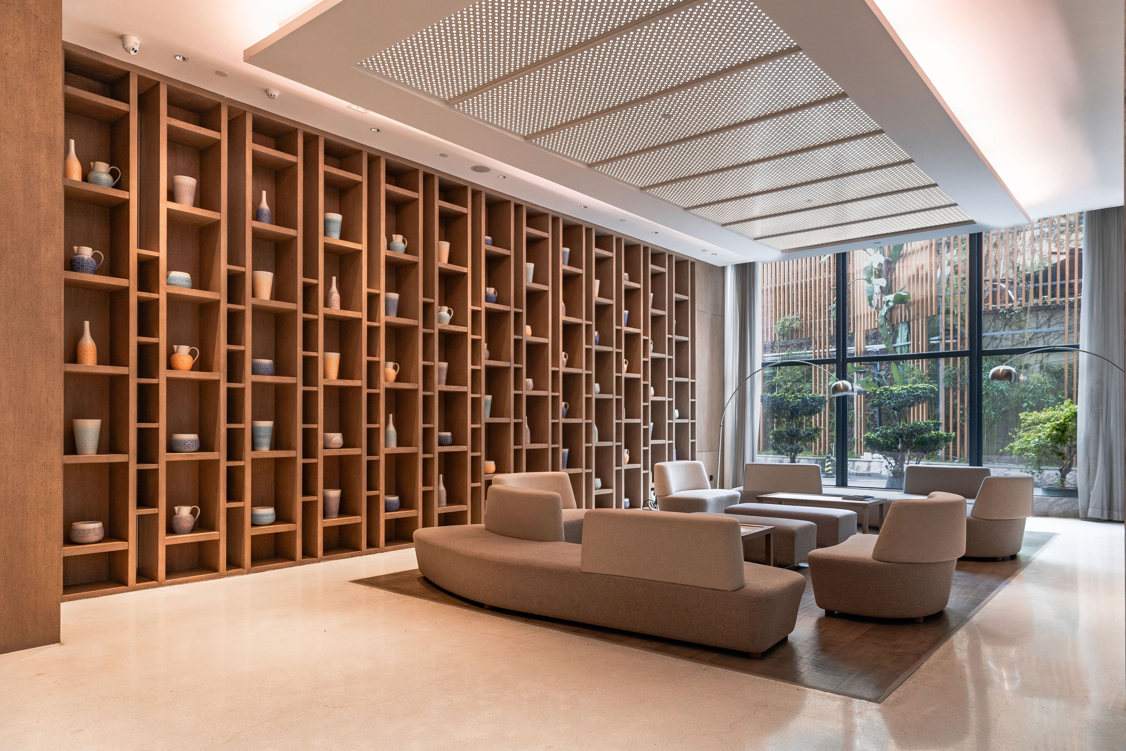A modern interior with a wall of shelves displaying various ceramic items, a seating area with sofas, and a large window offering a view of greenery.