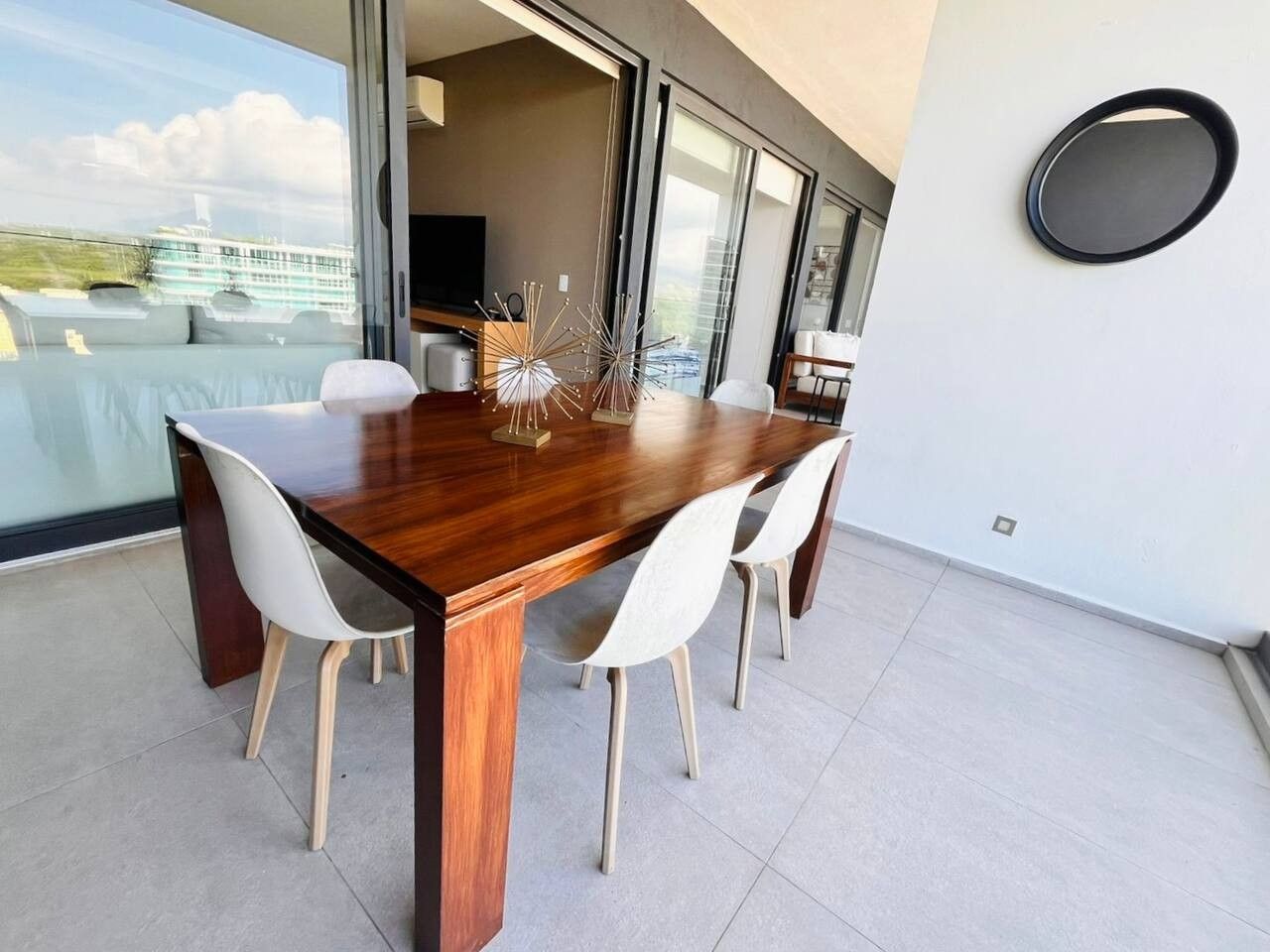Comedor con mesa de madera y cuatro sillas, un jarrón decorativo y vistas a un edificio a través de amplios ventanales.