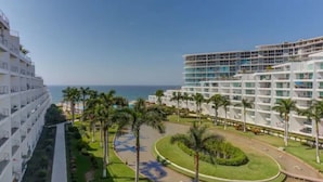Hotel com vários andares, um jardim bem cuidado com palmeiras e vista para o oceano.