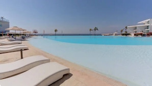 Uma área à beira da piscina com espreguiçadeiras e guarda-sóis, céu claro e um edifício ao fundo.