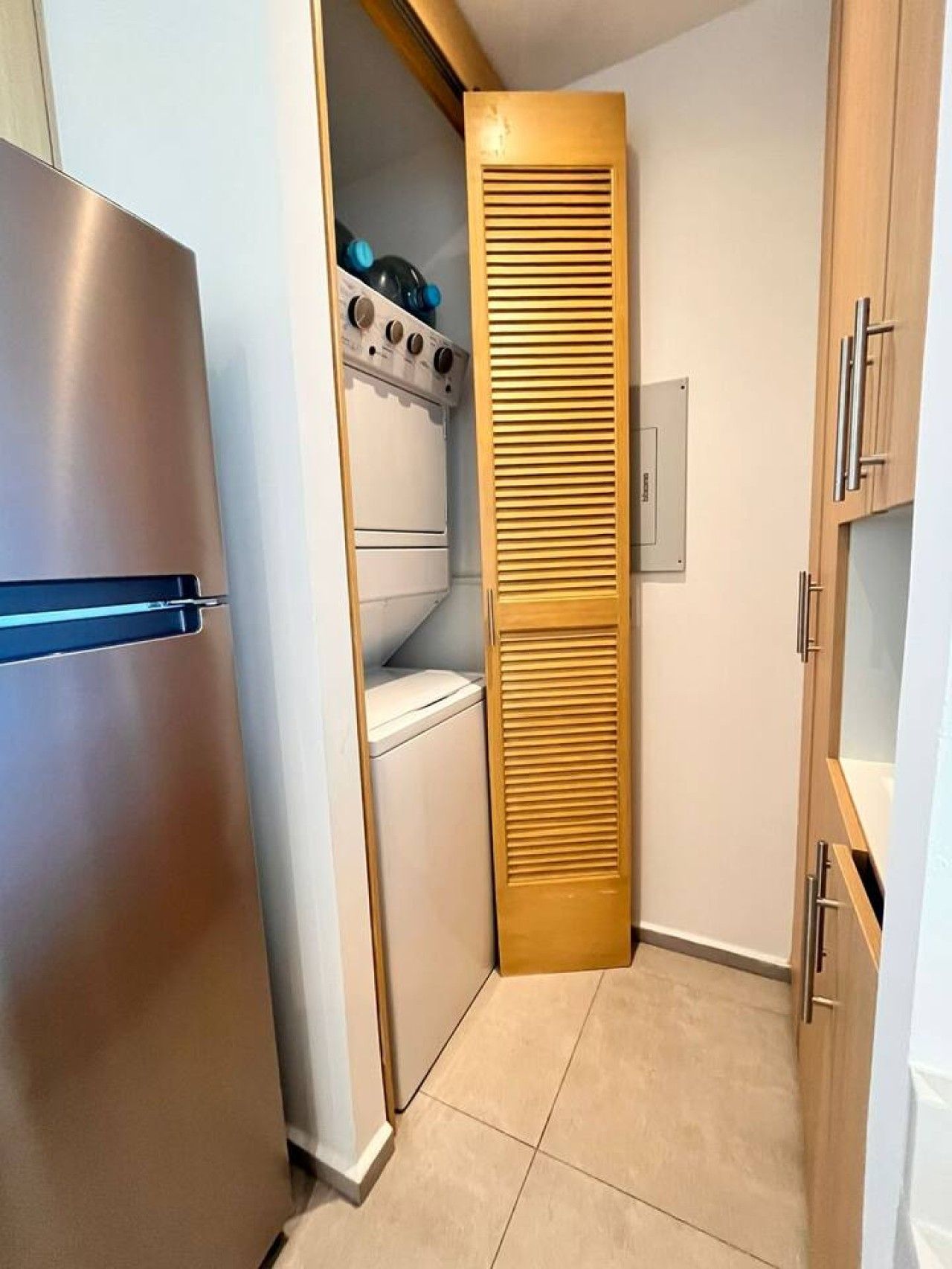 A compact kitchen with a stainless steel refrigerator, a built-in oven, and a wooden cabinet with a louvered door.