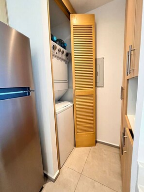Cocina compacta con refrigerador de acero inoxidable, horno integrado y mueble de madera con puerta persiana.
