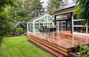 Ett modernt hus med ett inglasat uterum, en träterrass och en grillplats.