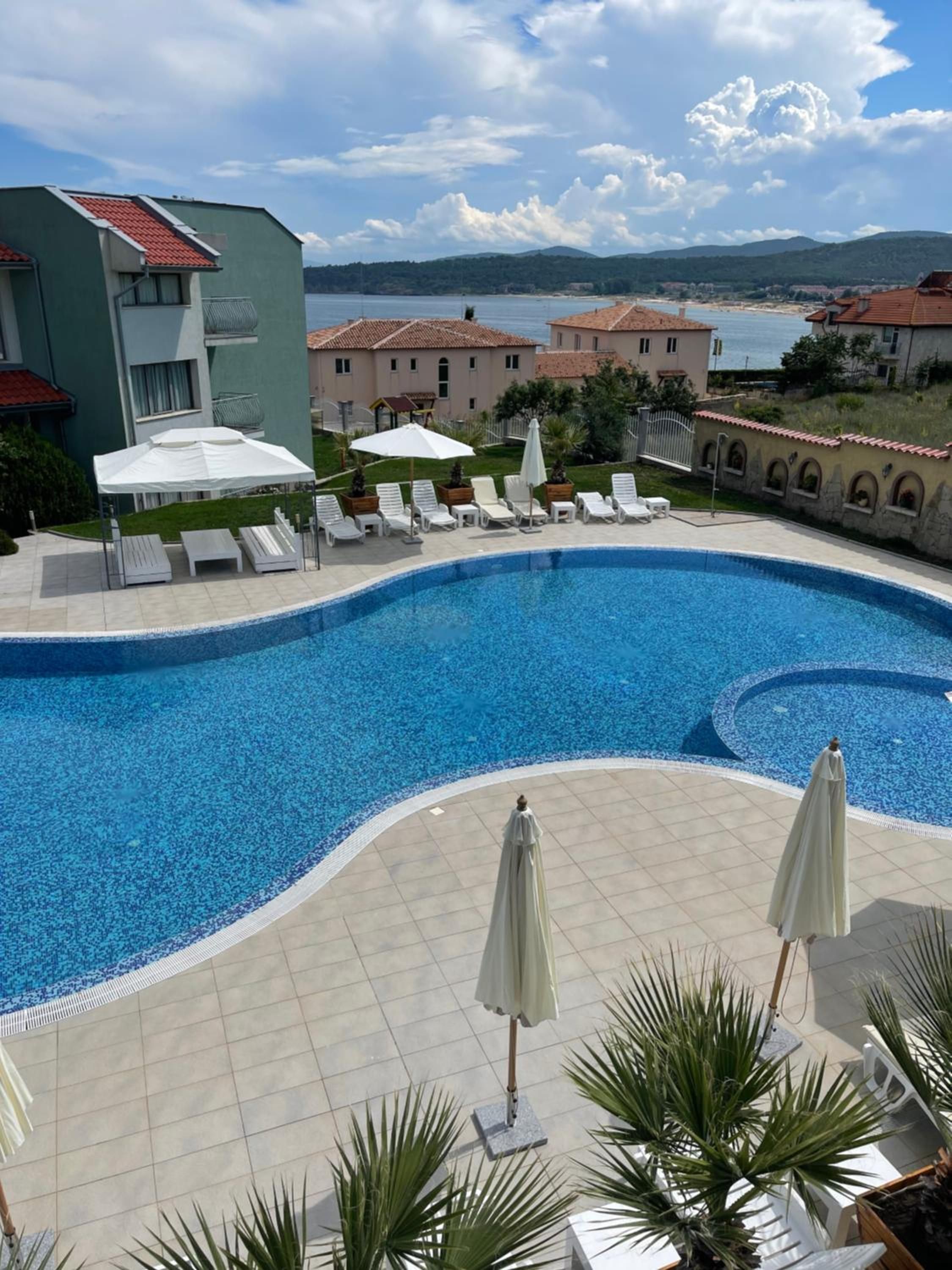 Piscina con sillas de descanso y sombrillas, rodeada de edificios y vista al mar.