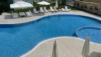 Piscina con sillas de descanso y sombrillas, rodeada de edificios y vista al mar.