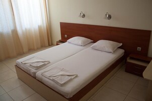 Cama doble con ropa de cama y toallas blancas, cabecera de madera, dos mesitas de noche y una ventana con cortinas.