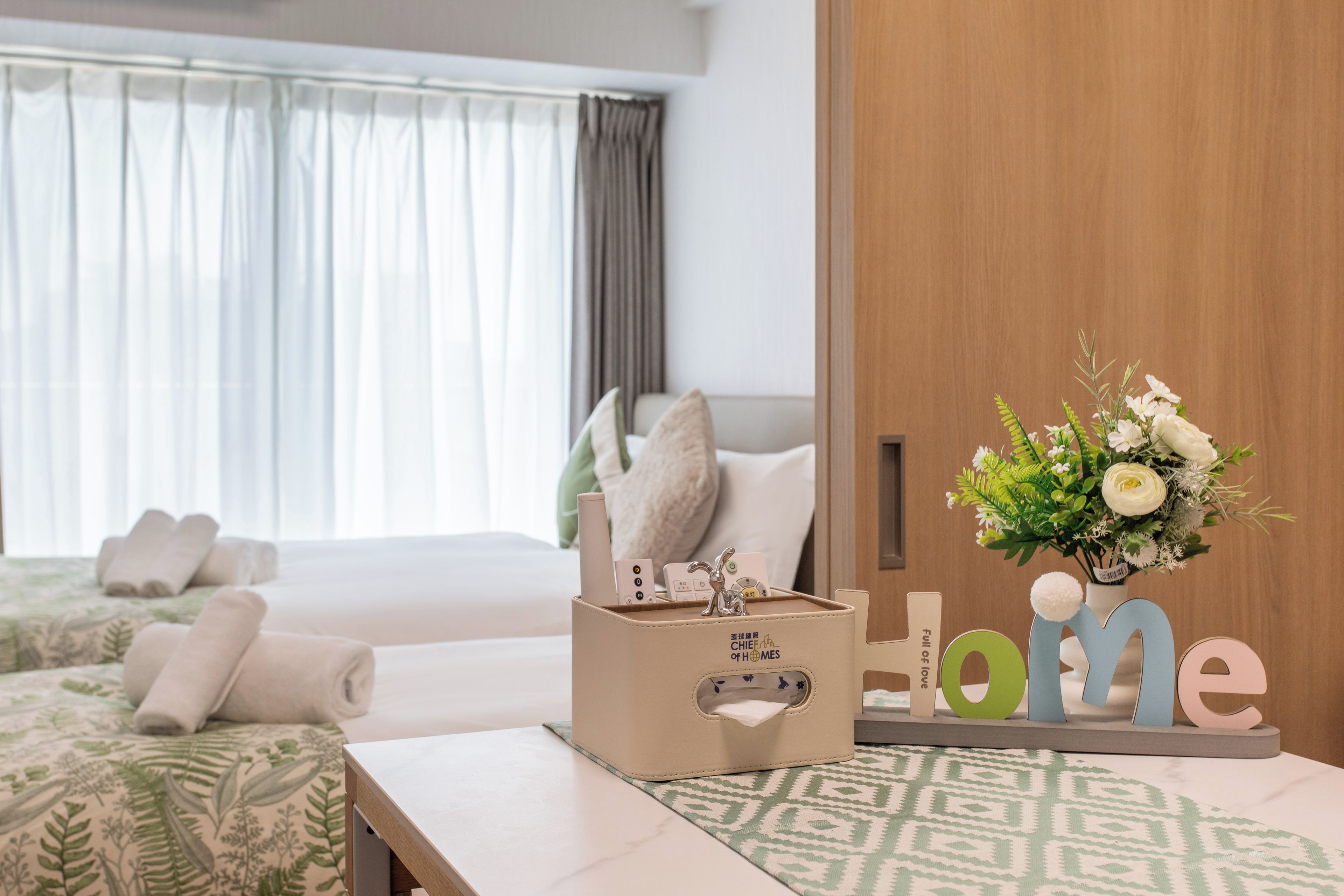 A hotel room with a bed, a bedside table with a tissue box, a decorative sign with letters, and a window with curtains.