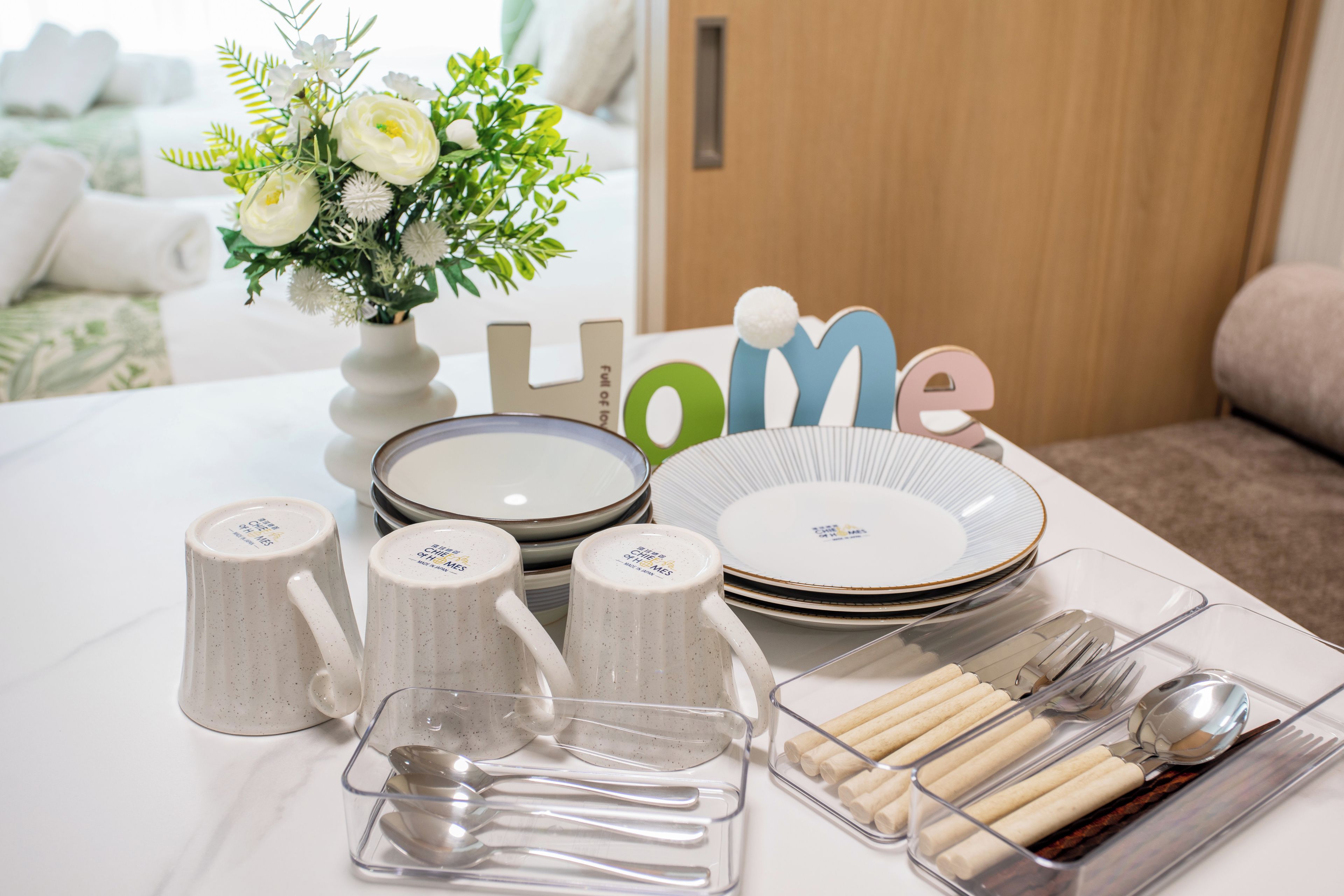 A table set with white ceramic mugs, plates, and cutlery, a vase with flowers, and a 'Home' decoration.