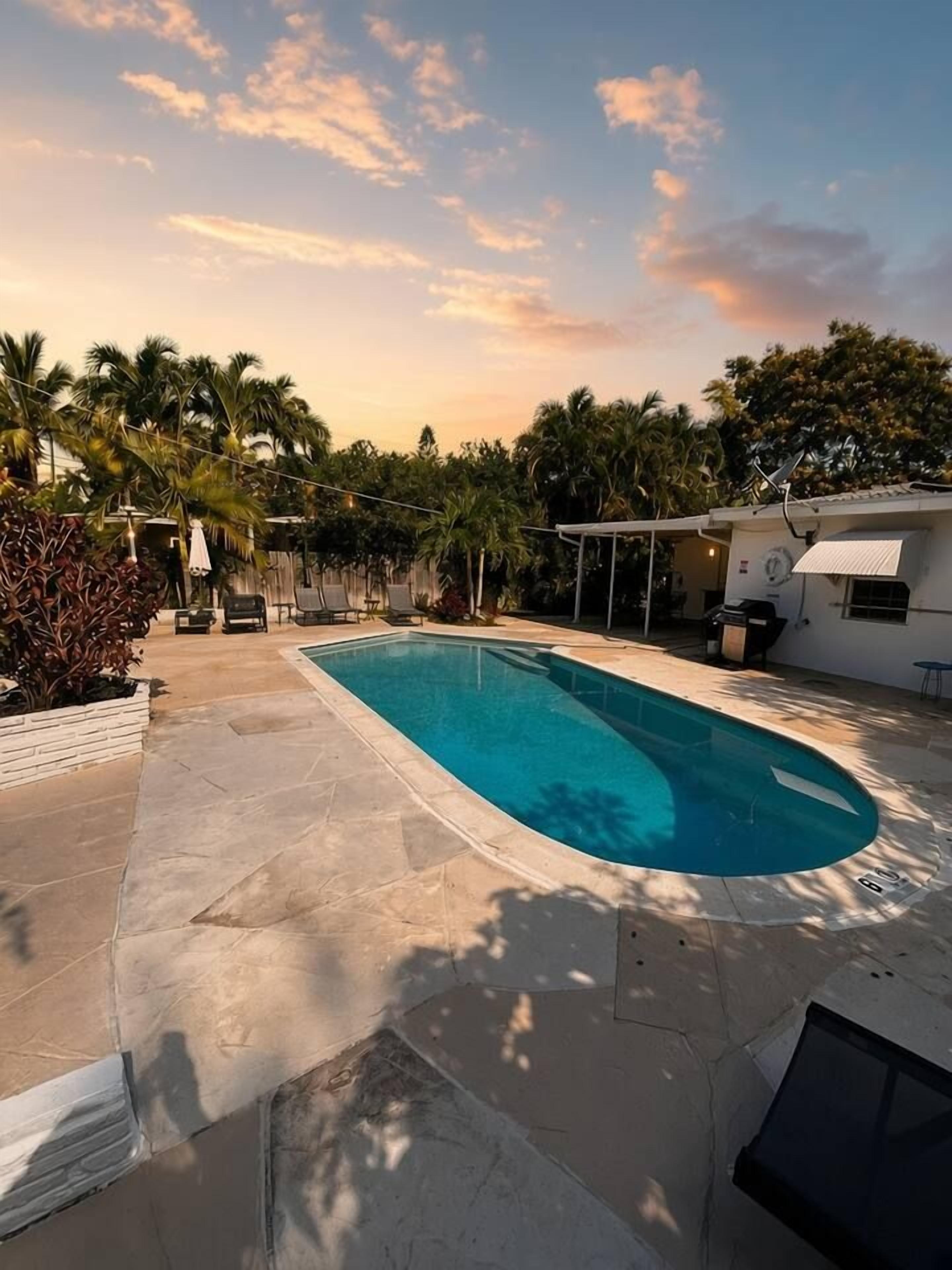 A swimming pool with a tiled deck, surrounded by palm trees and a covered patio area.