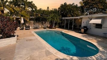 A swimming pool with a tiled deck, surrounded by palm trees and a covered patio area.