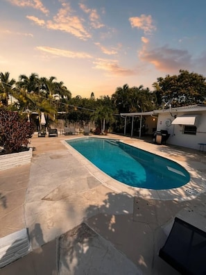 A swimming pool with a tiled deck, surrounded by palm trees and a covered patio area.