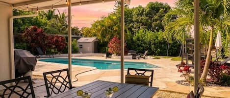 An outdoor patio with a pool, surrounded by greenery and a covered seating area.