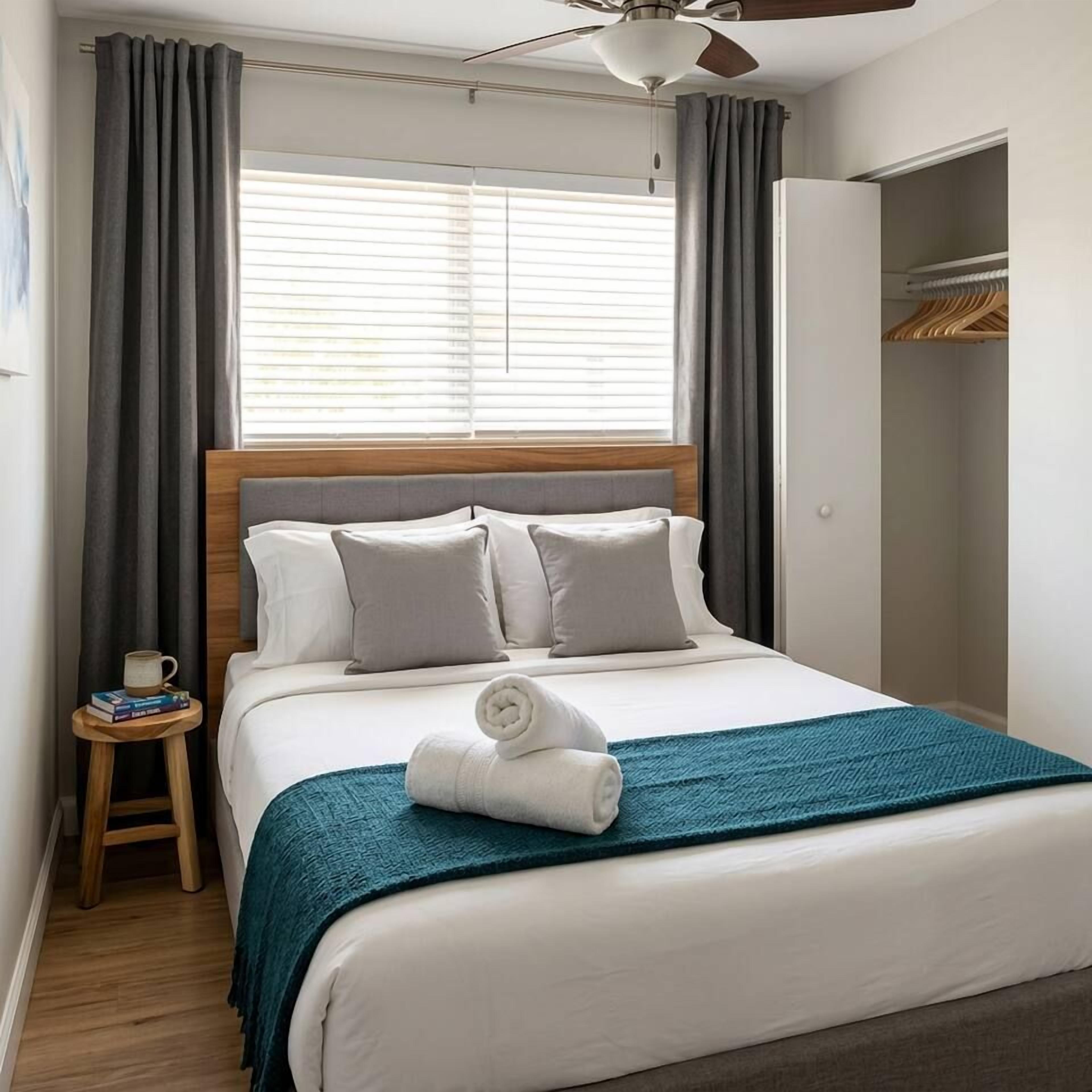 Une chambre d’hôtel avec un lit, une table de chevet sur laquelle se trouvent une tasse et des livres, un ventilateur de plafond et une armoire.