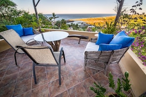 An outdoor patio with a view of the ocean, featuring two wicker chairs with blue cushions, a round table, and a tiled floor.