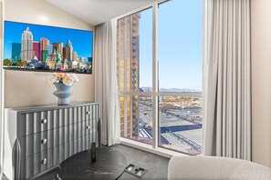 Une chambre d’hôtel dotée d’une grande fenêtre donnant sur le paysage urbain, d’une console grise sur laquelle est posé un vase de fleurs, et d’une télécommande sur le sol.