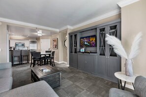 A modern living room with a grey sofa, a coffee table, a dining area with chairs, and a grey entertainment unit with a flat-screen TV. - Strip View Suite|On LV Strip w/Balcony/LazyRiver (Las Vegas)