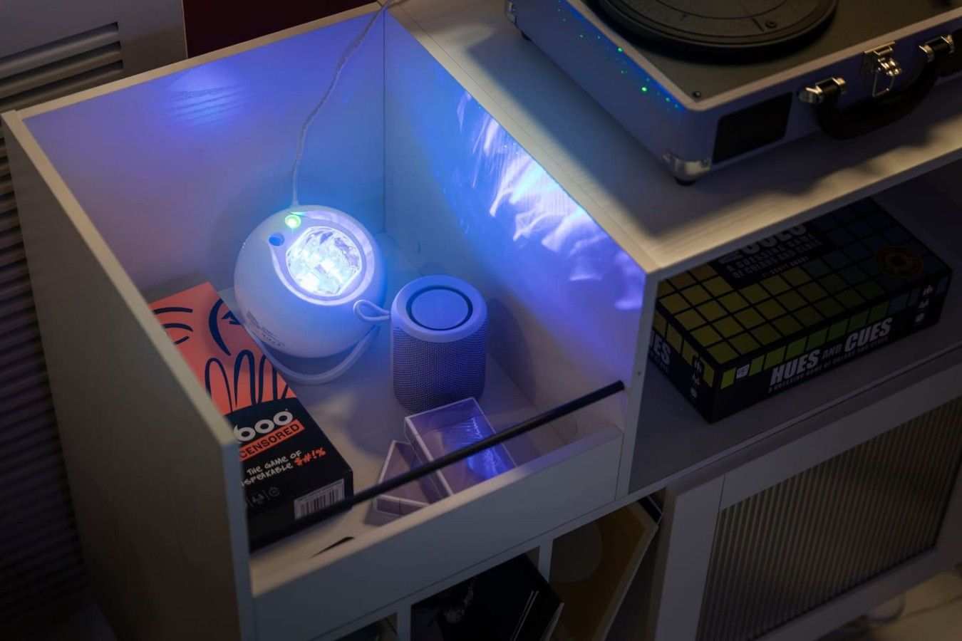 A room with a shelf containing a game box, a speaker, and a box of HUES AND CUES.
