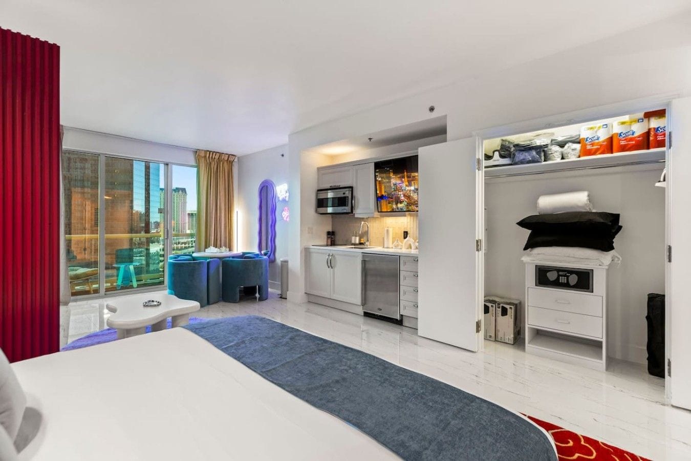 A hotel room with a bed, a desk, a kitchenette, and a view of the city.