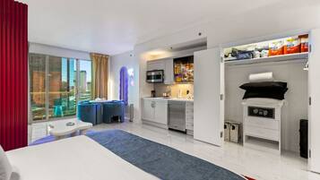 A hotel room with a bed, a desk, a kitchenette, and a view of the city.