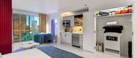 A hotel room with a bed, a desk, a kitchenette, and a view of the city.