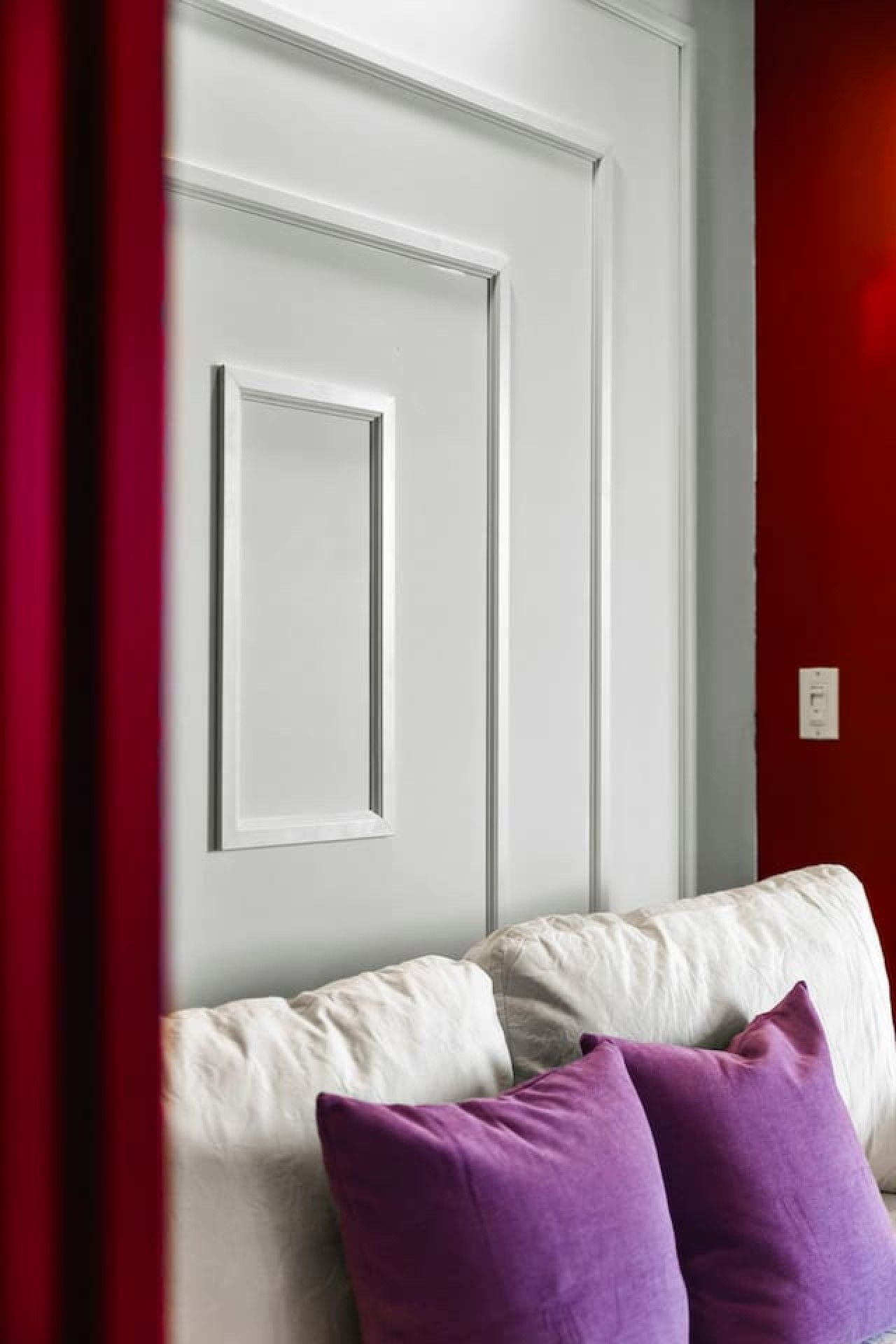 A white door with a simple design, a white wall, and a red wall with a light switch.