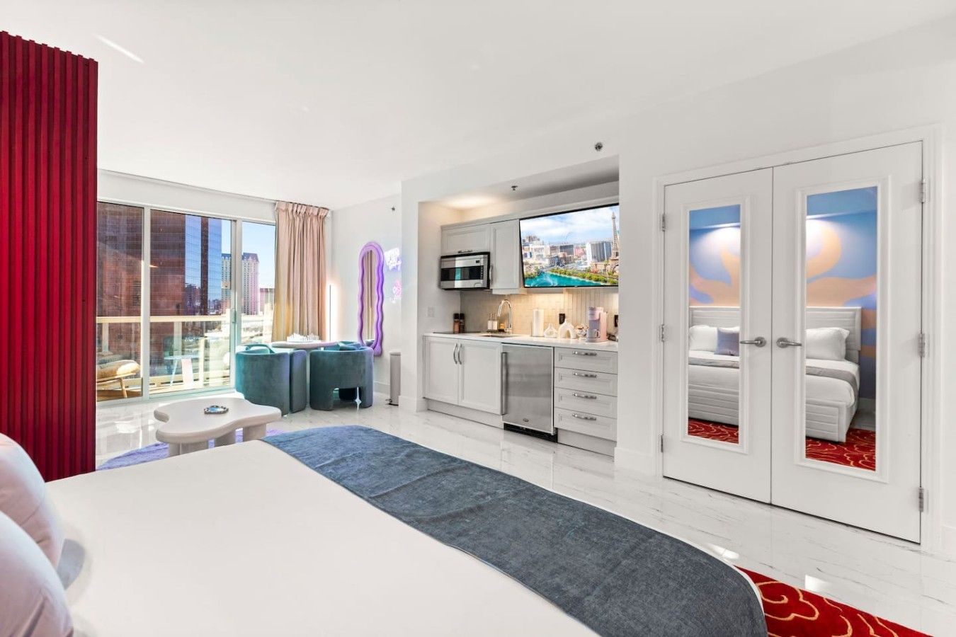 A hotel room with a bed, a TV, a kitchenette, and a large window with a city view.