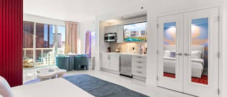 A hotel room with a bed, a TV, a kitchenette, and a large window with a city view.