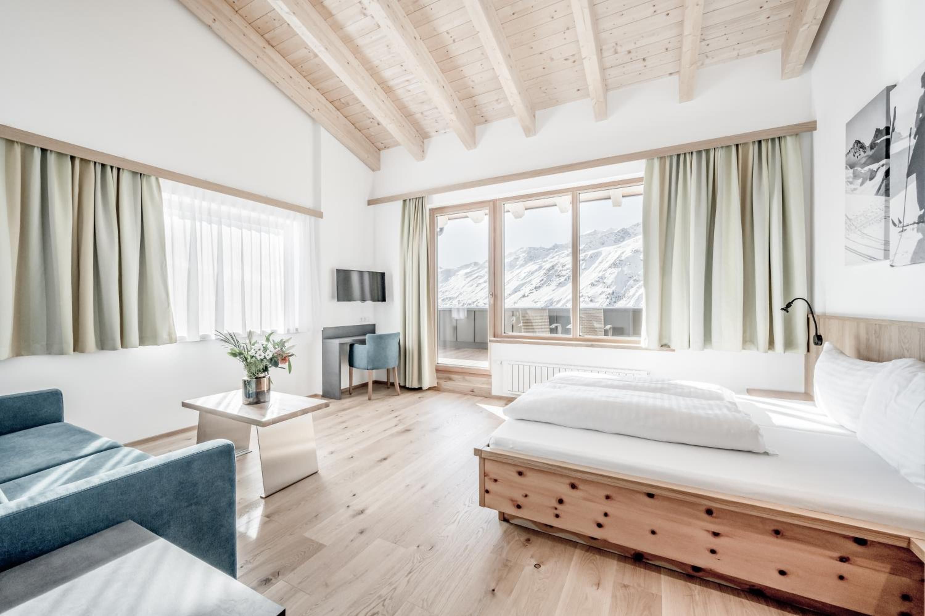 A modern hotel room with a wooden ceiling, a bed, a desk, a chair, and a view of mountains through large windows.