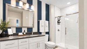 Un baño moderno con un espejo grande, muebles blancos, cabina de ducha y una pequeña planta en el tocador.
