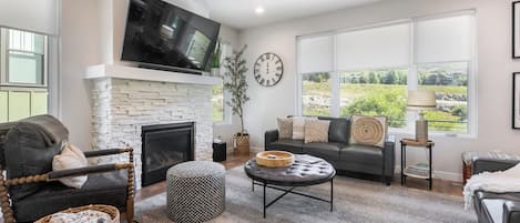 Una sala de estar con chimenea, televisor montado sobre ella, sofá, mesa de centro y una ventana grande con vista a follaje.