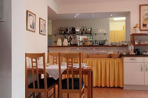 A dining area with wooden chairs and a table, a kitchen with shelves of bottles and kitchenware, and a yellow curtain. - Hotel Deisterblick (Bad Nenndorf)