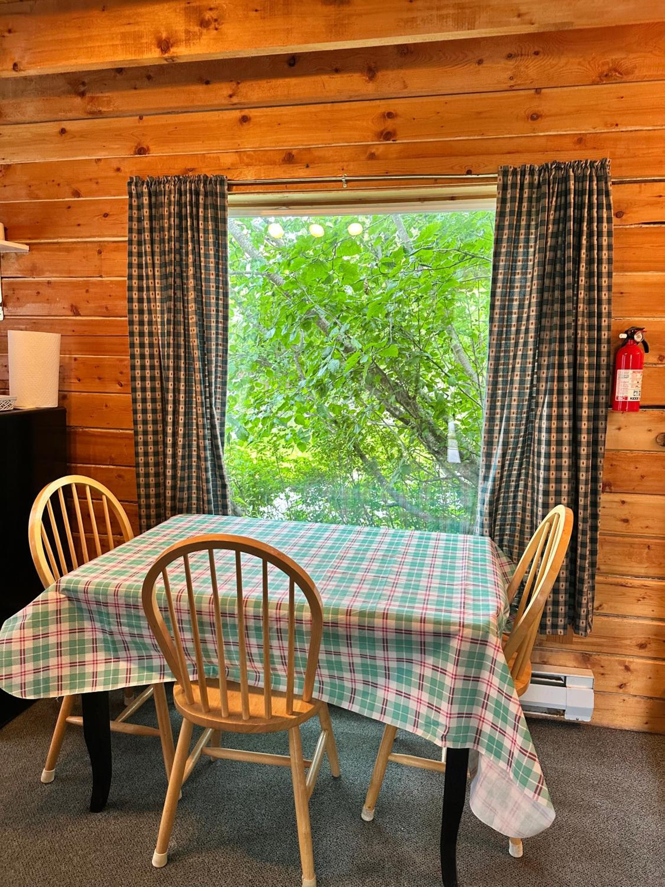 A wooden cabin interior with a table set for four, two chairs, a window with curtains, and a fire extinguisher.