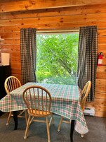 Interior de una cabaña de madera con una mesa dispuesta para cuatro personas, dos sillas, una ventana con cortinas y un extintor.