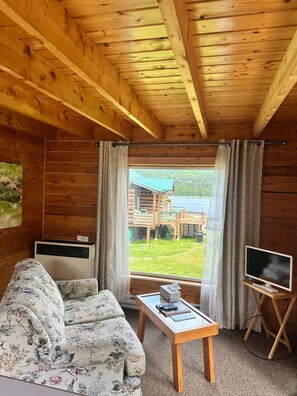 Interior de una cabaña de madera con sofá, una mesa, un televisor sobre un mueble bajo y una ventana con cortinas.