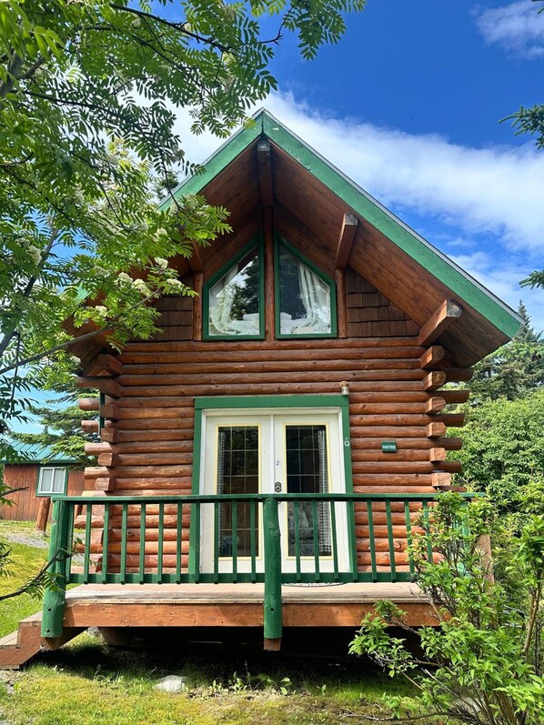 Una cabaña de troncos con un porche verde y una puerta blanca, rodeada de árboles.