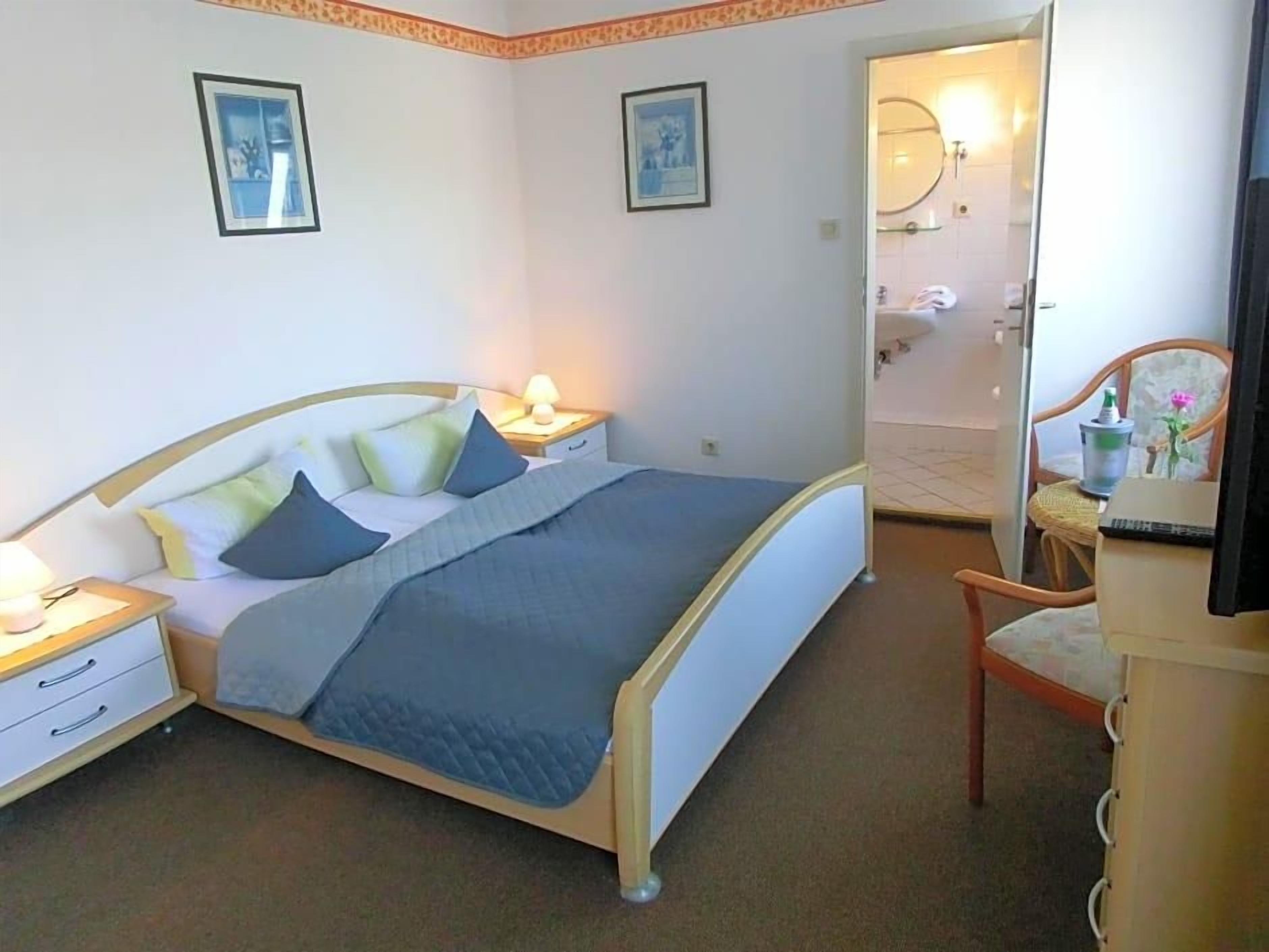 Ein Schlafzimmer mit einem Bett, Nachttischen, einem Sessel und einem durchgehenden Tür angedeuteten Badezimmer.