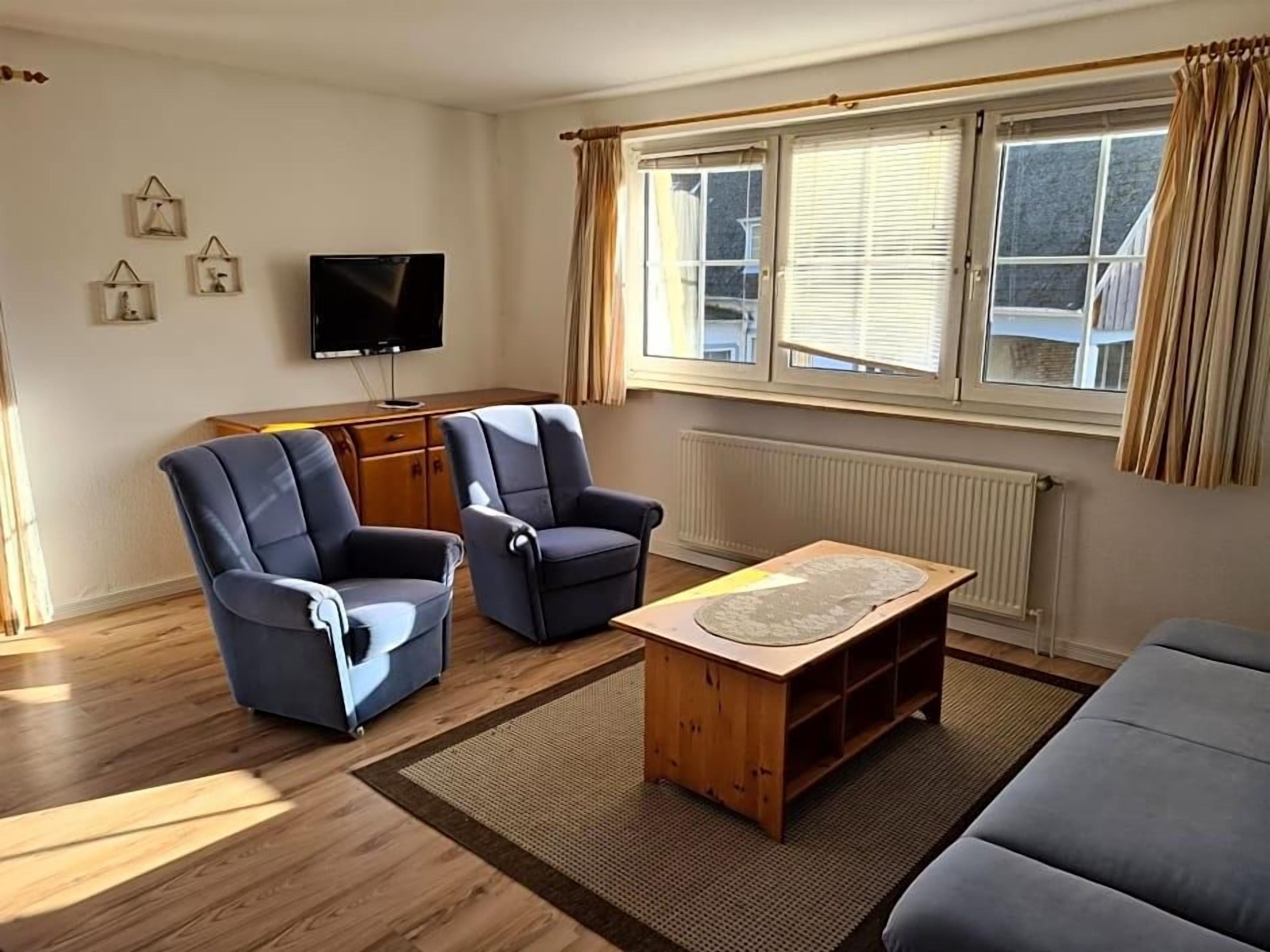 Ein Wohnzimmer mit zwei Sesseln, einem Holztisch, einem Fernseher und einem Fenster mit Vorhängen.