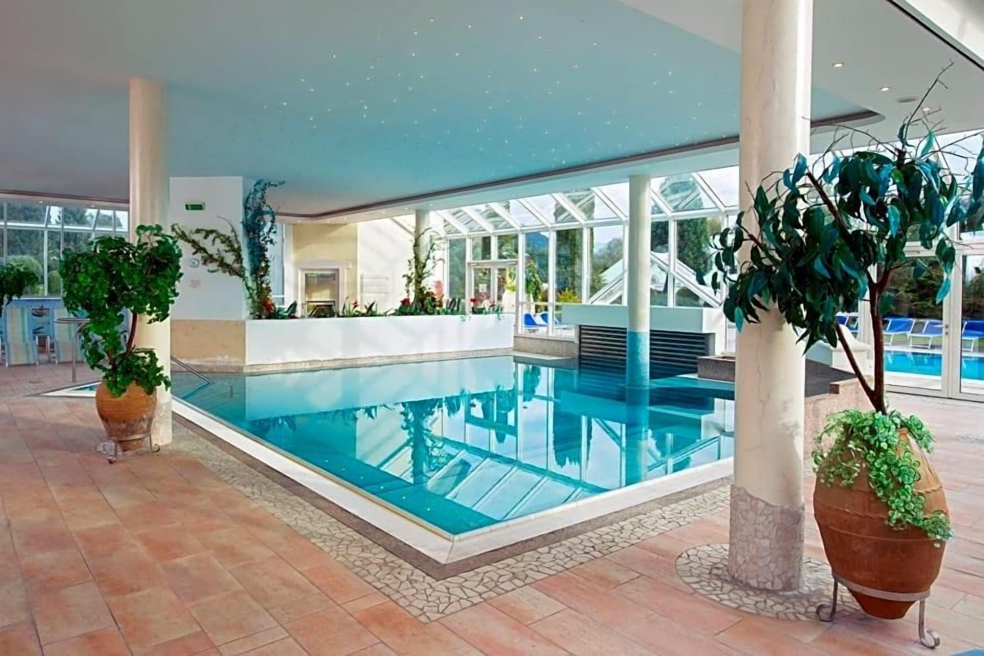 An indoor swimming pool with clear glass walls, surrounded by potted plants and a tiled floor.