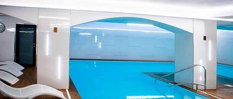 Ein moderner, überdachter Schwimmbereich mit klarem, blauem Wasser, einer Glaskonstruktion und einem Holzboden.