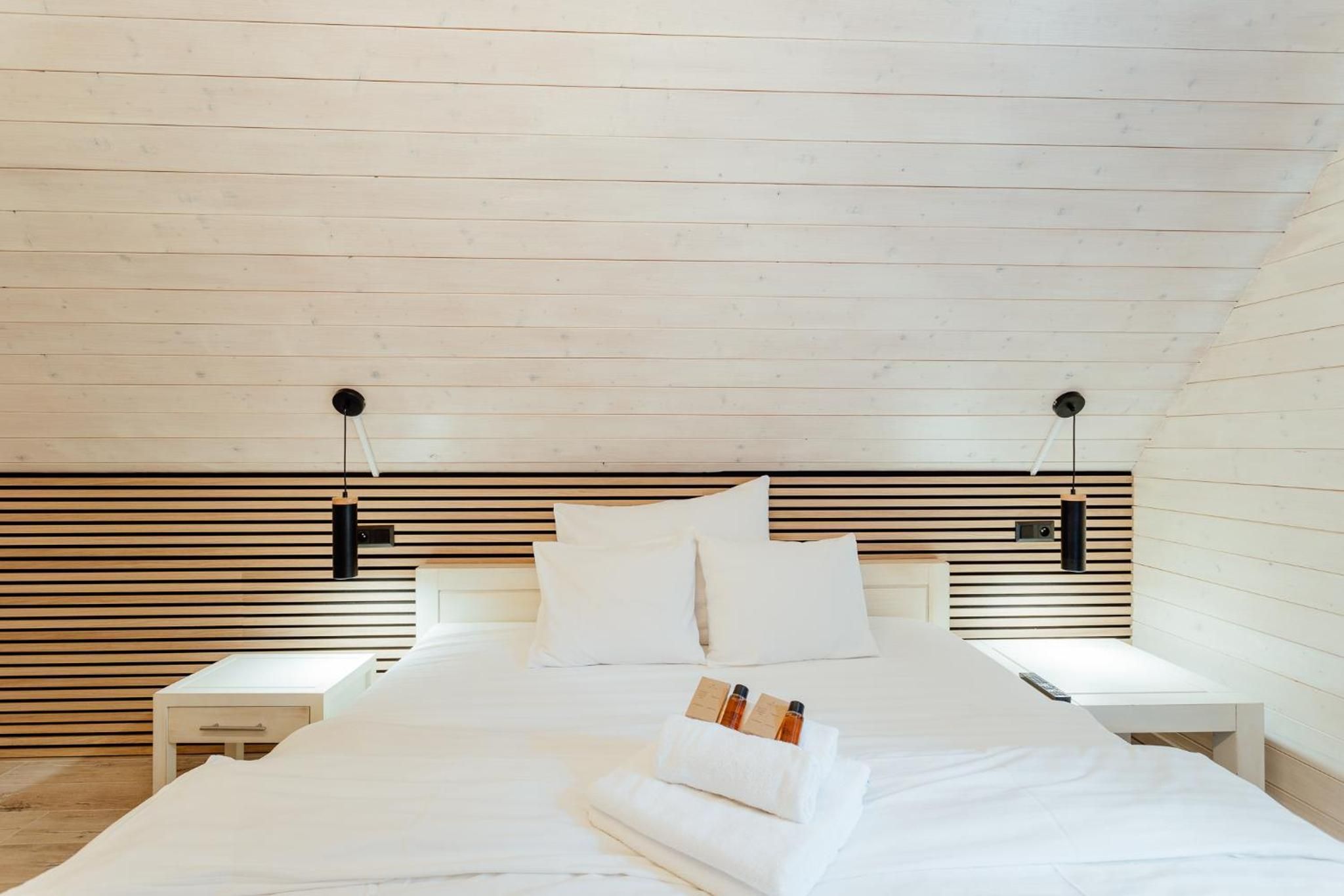 Un dormitorio con techo inclinado, cabecera de madera, ropa de cama blanca, dos mesitas de noche y una mesilla.