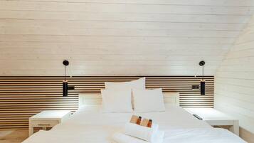 Un dormitorio con techo inclinado, cabecera de madera, ropa de cama blanca, dos mesitas de noche y una mesilla.