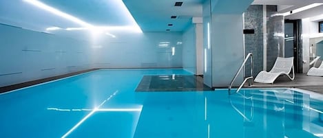 A modern indoor swimming pool with clear water, a sunlit ceiling, and lounge chairs by the poolside.