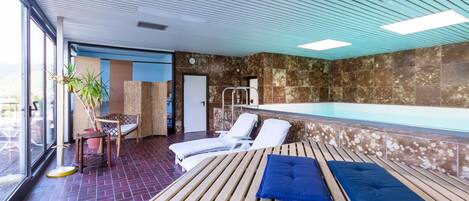 Un espace piscine intérieure avec des chaises longues, un banc en bois et une vue sur l’extérieur.